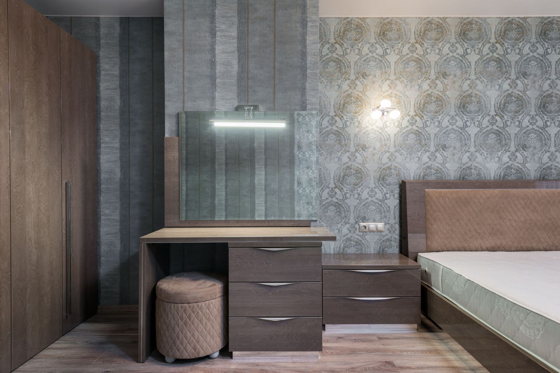 Wooden cupboard with mirror and pouf placed near wardrobe and bed with mattress at wall with wallpaper in spacious bedroom